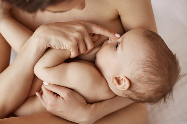 Young tender nude mother breastfeeding hugging her newborn baby sitting in bed at morning.