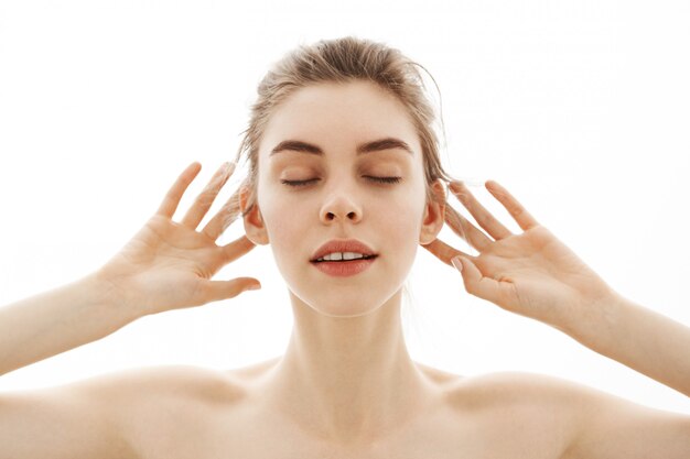 Young tender nude woman with bun posing over white background. Closed eyes.