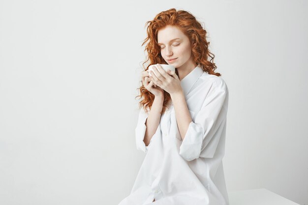 Young tender redhead girl in shirt smiling holding cup sitting on table Closed eyes.