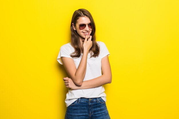 Молодая думающая дама в белой футболке и синих джинсах остается на желтом фоне студии