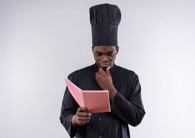 Молодой вдумчивый афро-американский повар в униформе шеф-повара держит и смотрит в блокнот на белом с копией пространства