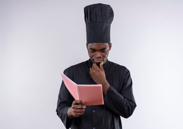 Young thoughtful afro-american cook in chef uniform holds and looks at notebook on white  with copy space