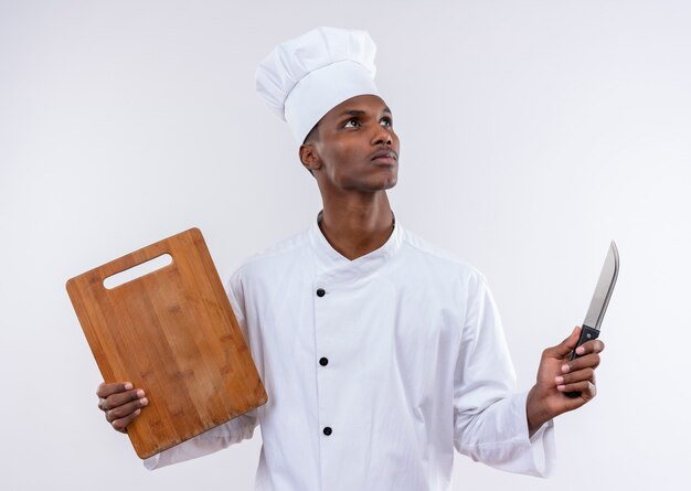 요리사 제복을 입은 젊은 사려 깊은 아프리카 계 미국인 요리사는 흰 벽에 고립 된 나무 주방 책상과 칼을 보유하고 있습니다.