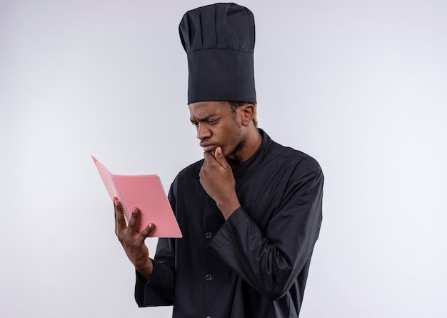 Young thoughtful afro-american cook in chef uniform looks at notebook isolated on white wall 