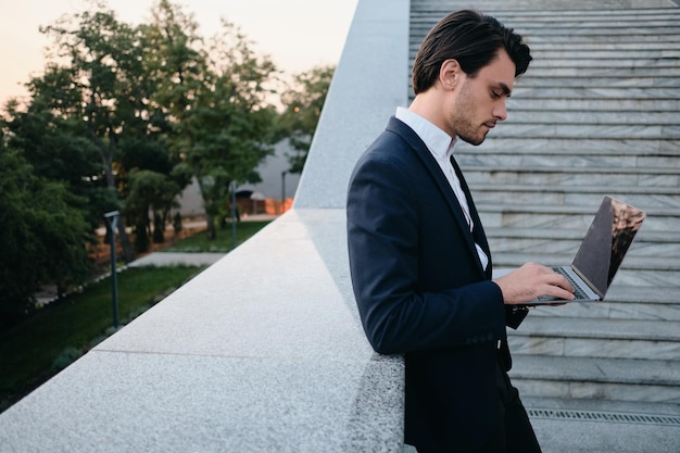 Young thoughtful bearded brunette man in white shirt and classic suit standing on street alone with laptop in hands