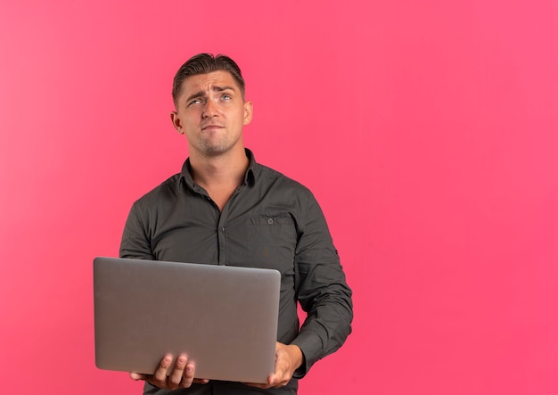Young thoughtful blonde handsome man holds laptop and looks up isolated on pink background with copy space