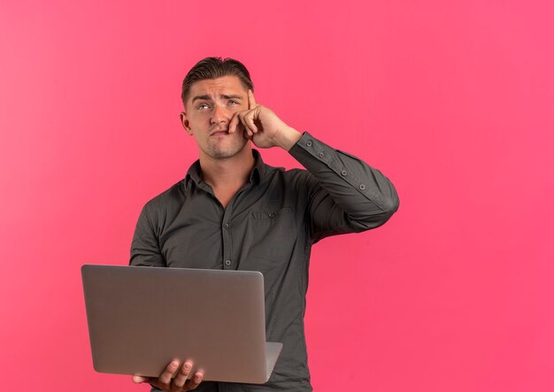 Young thoughtful blonde handsome man holds laptop and puts hand on face looking up isolated on pink background with copy space
