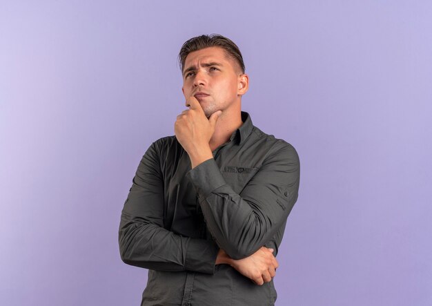 Young thoughtful blonde handsome man puts hand on chin and looks up isolated on violet background with copy space