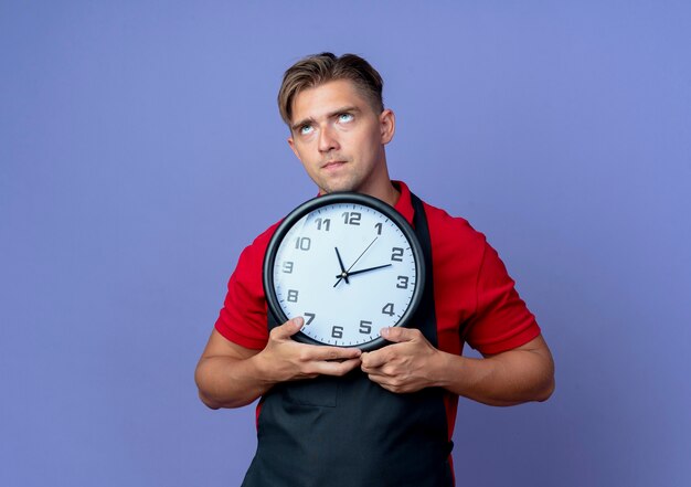 Молодой вдумчивый светловолосый парикмахер в униформе держит часы, глядя вверх изолированно на фиолетовом пространстве с копией пространства