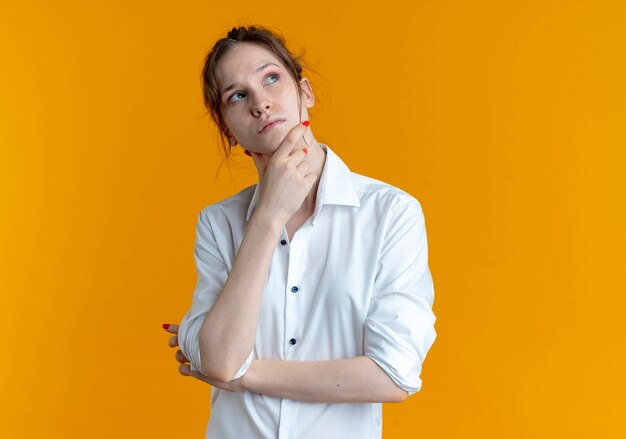 Young thoughtful blonde russian girl looks up holding chin isolated on orange space with copy space