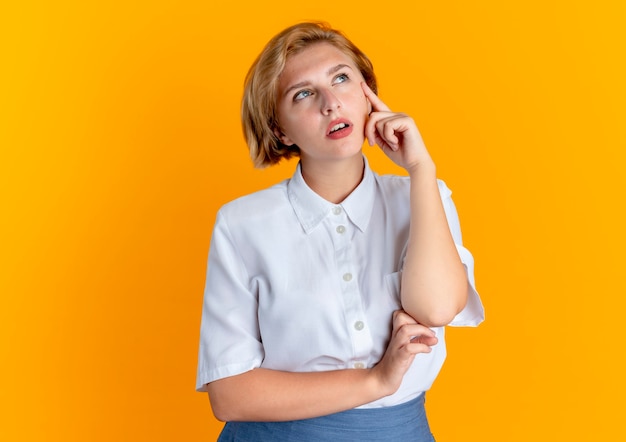 Young thoughtful blonde russian girl looks up and puts hand on face isolated on orange background with copy space