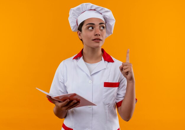 Young thoughtful caucasian cook girl in chef uniform holds notebook and points up isolated on orange wall 