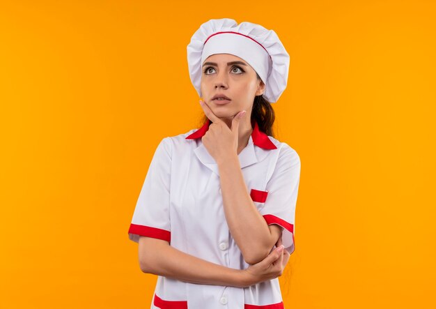 Young thoughtful caucasian cook girl in chef uniform puts hand on chin isolated on orange wall with copy space