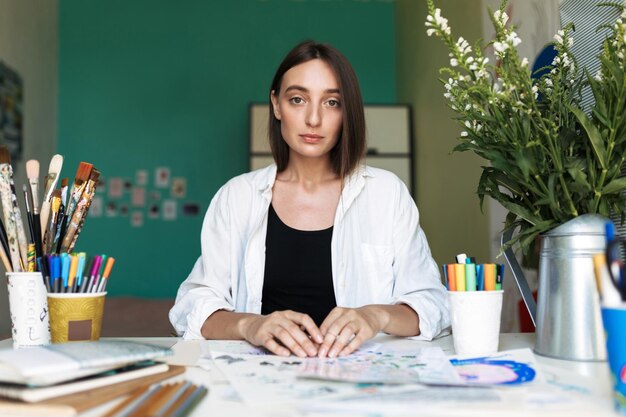 Молодая задумчивая девушка с темными волосами сидит за столом с картинками, мечтательно глядя в камеру, рисуя дома