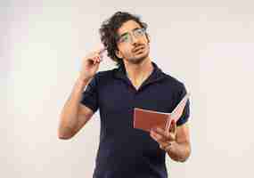 Free photo young thoughtful man in black shirt with optical glasses holds notebook and puts pen on head isolated on white wall