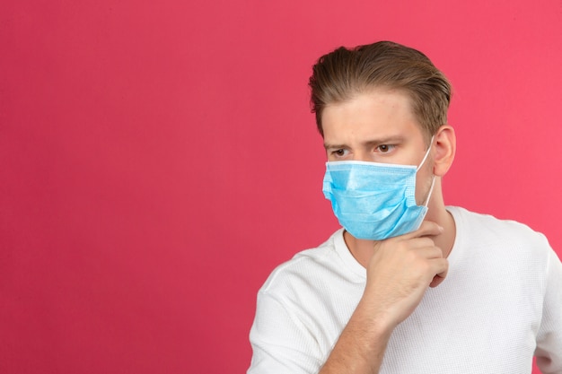Young thoughtful man in medical protective mask looking away and keeping hand on chin while standing over isolated pink background