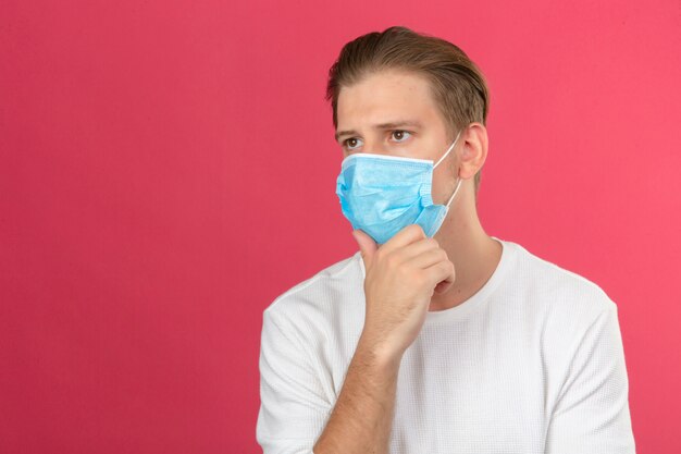 Young thoughtful man in medical protective mask touching chin and thinking over isolated pink background