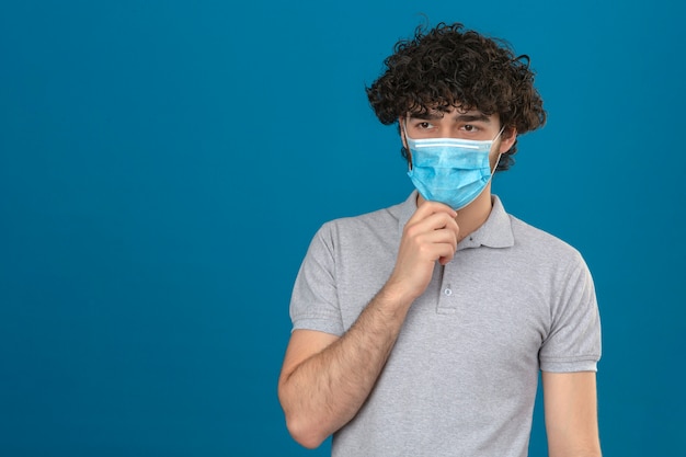 Young thoughtful man in medical protective mask with hand on chin pensive look standing over isolated blue background