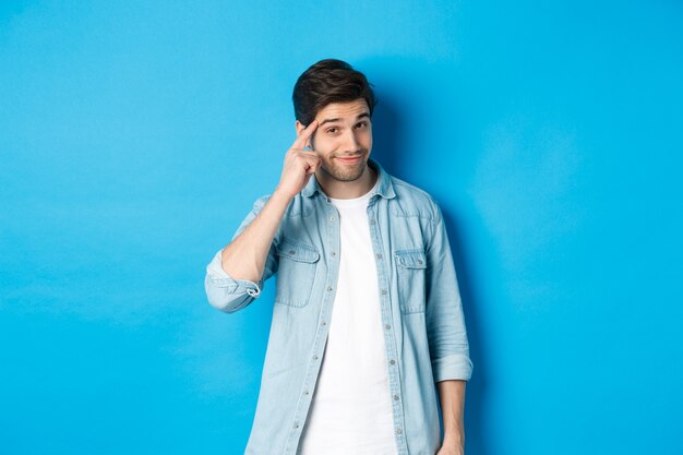 Young thoughtful man pointing at head, asking to think about it, giving a hint, standing in casual clothes over blue background.