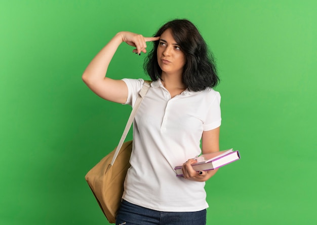 Young thoughtful pretty caucasian schoolgirl wearing back bag puts finger on head holding books on green  with copy space