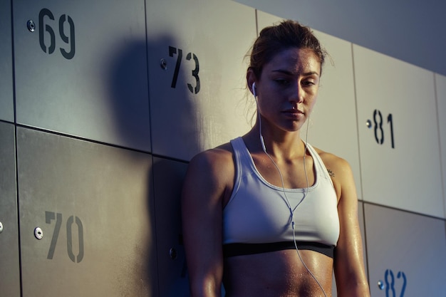 Free photo young thoughtful sportswoman standing in gym's dressing room and listening music on earphone after sports training