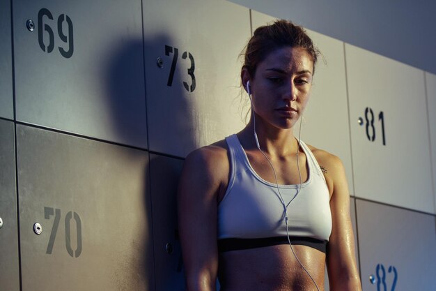 Young thoughtful sportswoman standing in gym's dressing room and listening music on earphone after sports training