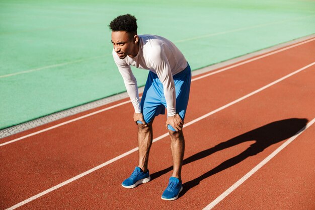 Молодой уставший афроамериканский спортсмен отдыхает на ипподроме после пробежки на городском стадионе
