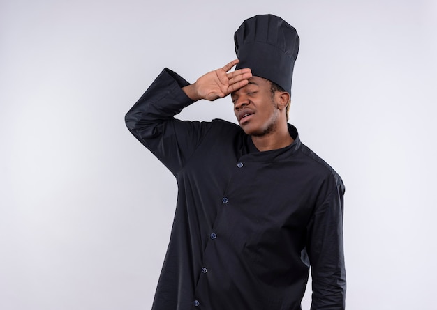 Young tired afro-american cook in chef uniform holds head with hand isolated on white wall 