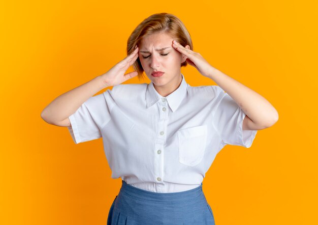 Young tired blonde russian girl holds head isolated on orange background with copy space