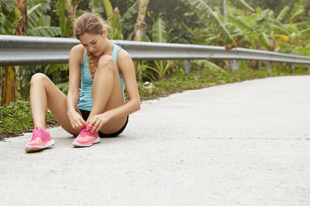 Young tired Caucasian woman runner lacing her pink running shoes, sitting on road in tropical forest having small break while jogging outdoors.