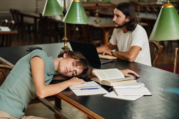 Молодая уставшая студентка спит на столе с книгами во время учебы в библиотеке университета
