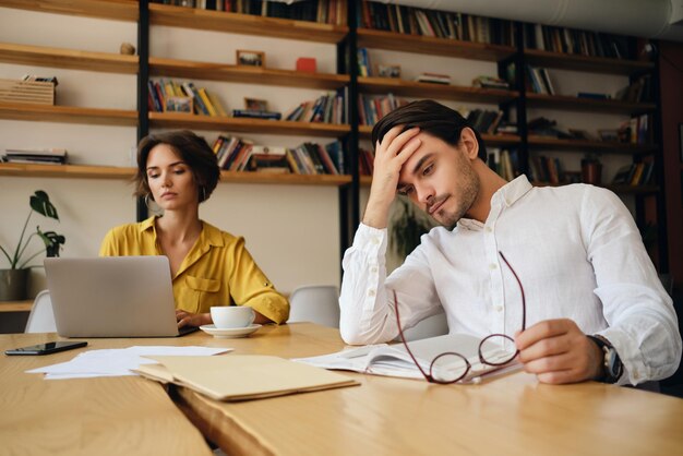 Young tired man sitting at the table with notepad and eyeglasses while thoughtfully holding hand near head at work in office with colleague on background