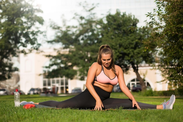 Молодая уставшая женщина больших размеров в розовом спортивном топе и леггинсах пытается сидеть на шпагате на коврике для йоги, проводя время в городском парке