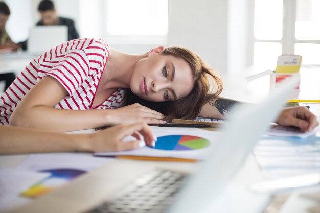現代のオフィスで時間を過ごしている間、夢のように脇を見て机の上に横たわっている縞模様のTシャツの若い疲れた女性