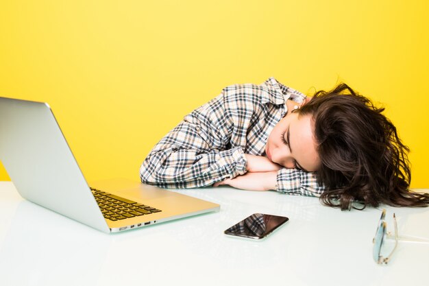 노란색 벽에 고립 된 노트북에서 일하는 젊은 피곤 된 여자