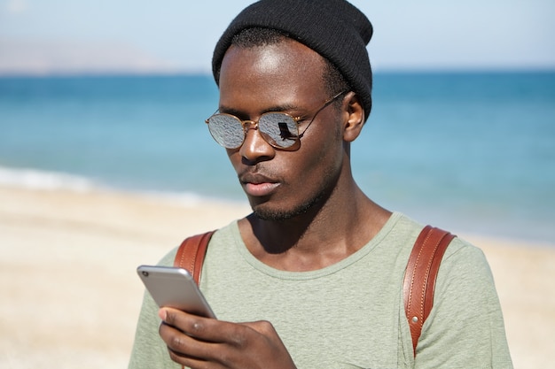 молодой турист, одетый в стильную одежду, набирает текстовое сообщение на мобильном телефоне
