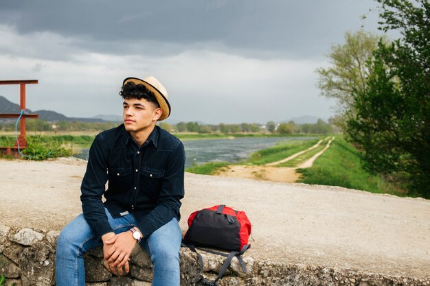 Шляпа молодого туриста нося распологая с рюкзаком на мосте