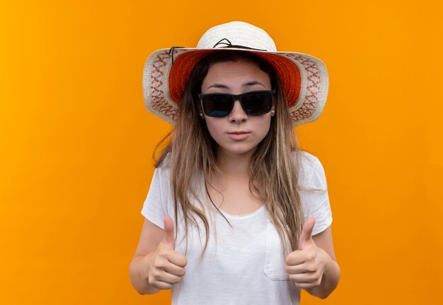 Молодая туристка в белой футболке, летней шляпе и черных очках показывает палец вверх, стоя над оранжевой стеной