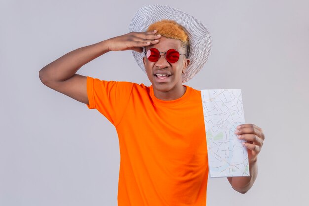 Young traveler boy in summer hat wearing orange t-shirt holding map looking confused emotional and worried standing over white wall