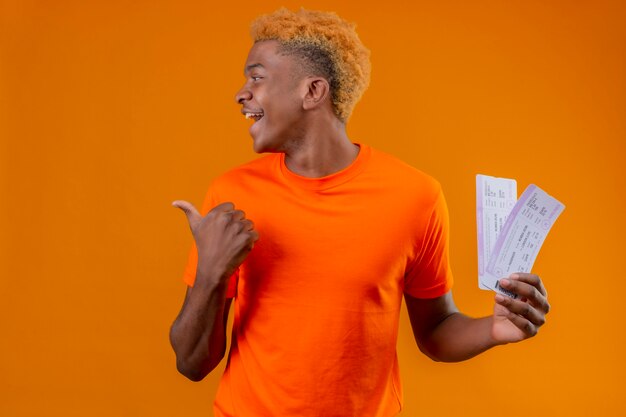 Young traveler boy wearing orange t-shirt holding airplane tickets looking aside smiling cheerfully pointing with thumb to the side standing over orange wall