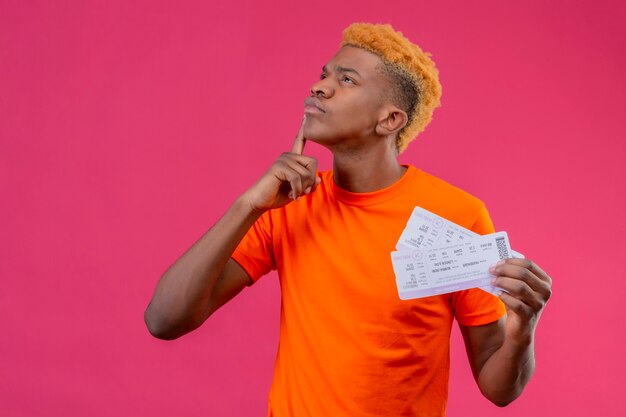 Молодой путешественник мальчик в оранжевой футболке держит билеты на самолет, глядя вверх с пальцем на подбородке с задумчивым выражением лица, думая, стоя над розовой стеной