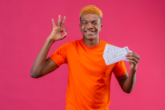 Young traveler boy wearing orange t-shirt holding airplane tickets smiling cheerfully doing ok sign standing over pink wall