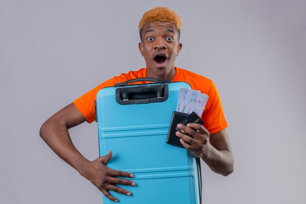 young traveler boy wearing orange t-shirt holding suitcase and air tickets surprised and amazed happy standing over white wall