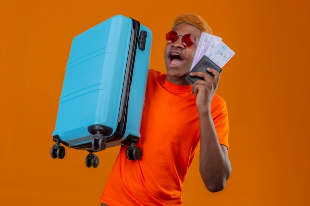 Young traveler boy wearing orange t-shirt holding suitcase and airplane tickets looking amazed and surprised standing over orange wall