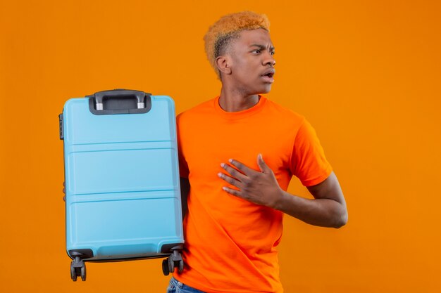 Young traveler boy wearing orange t-shirt holding suitcase looking aside confused and surprised touching his head standing over orange wall