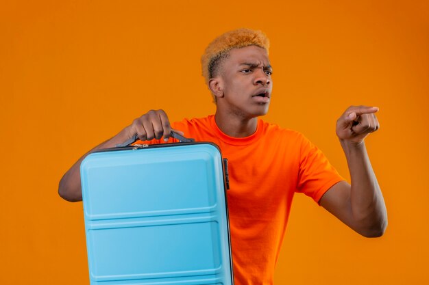 Young traveler boy wearing orange t-shirt holding suitcase looking aside pointing with finger to something intrigued standing over orange wall