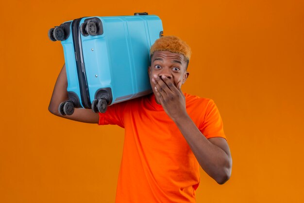 オレンジ色の壁の上に立って手で驚いて驚いて覆っている口を探しているスーツケースを持ってオレンジ色のtシャツを着ている若い旅行者少年