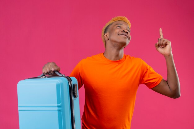 Young traveler boy wearing orange t-shirt holding suitcase pointing with finger up and looking up smiling standing over pink wall