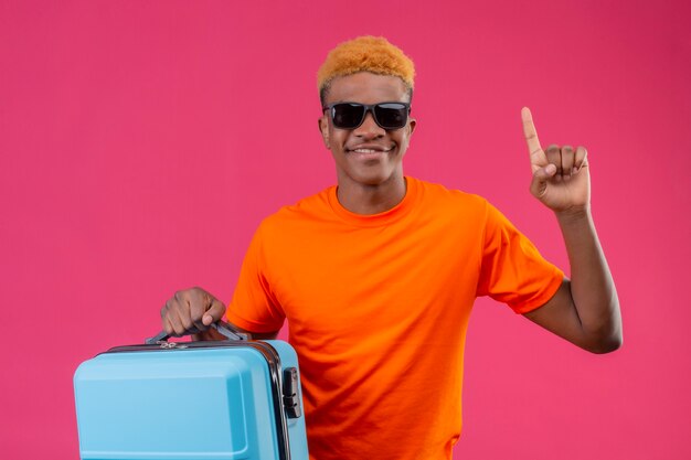 Young traveler boy wearing orange t-shirt holding suitcase pointing with finger up and smiling standing over pink wall