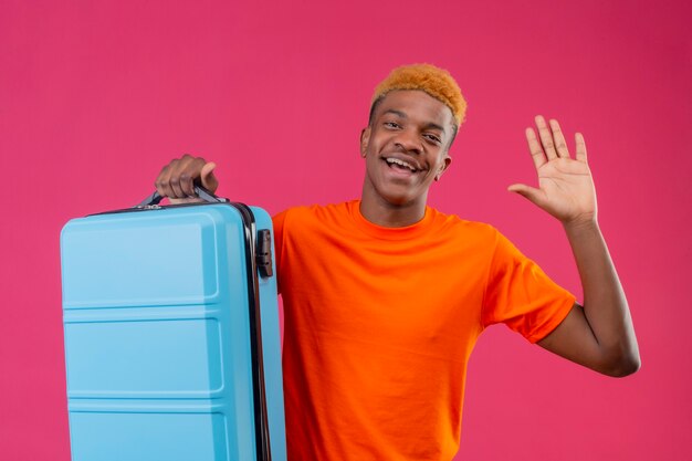 Young traveler boy wearing orange t-shirt holding suitcase smiling cheerfully waving with hand standing over pink wall